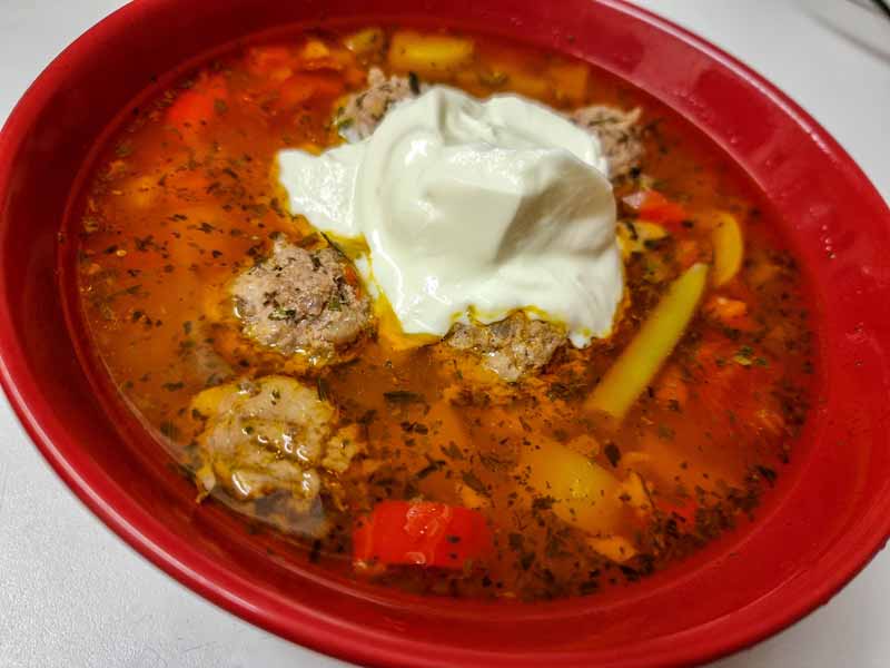 A red bowl with a red soup in it. You can see the meatballs half submerged in the soup. In the middle of the soup there is a dollop of white sour cream