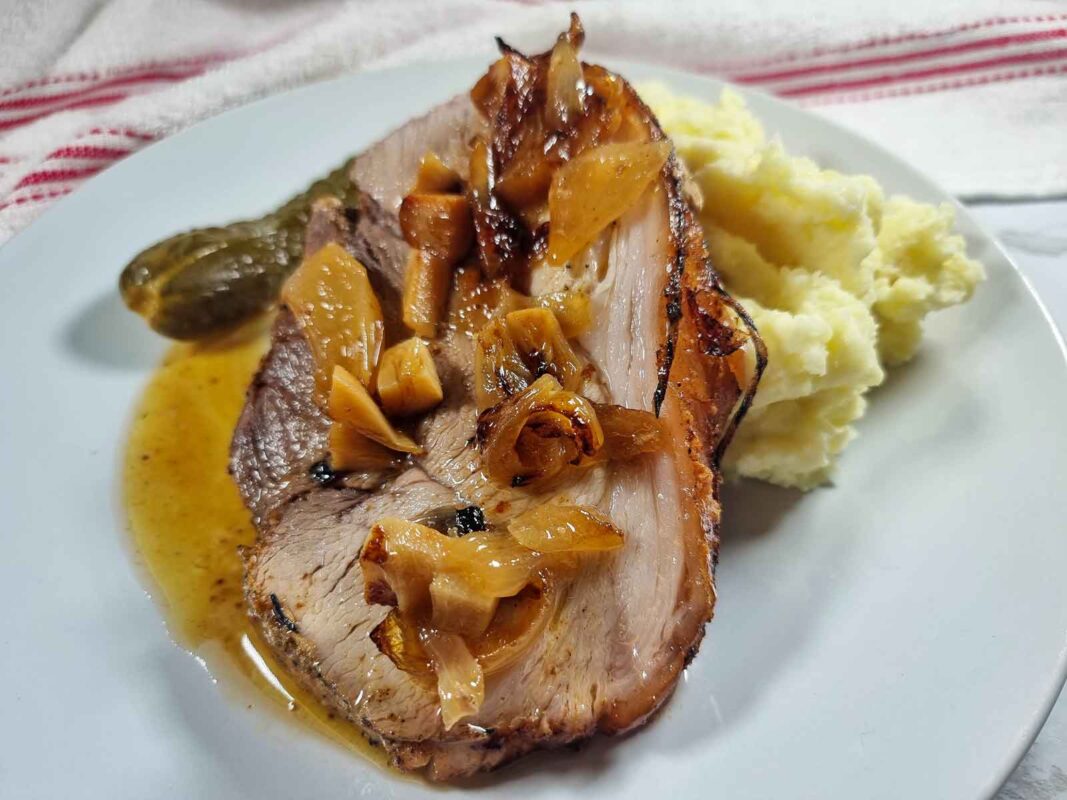 A slice of the roasted pork leg joint topped with sauce and roast garlic and onions, resting on mashed potatoes. In the back you can see a pickle. There is a light brown sauce on the white plate as well.