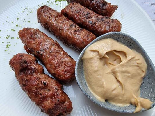 Small skinless sausages grilled, on a white plate, next to a small grey dish filled with mustard