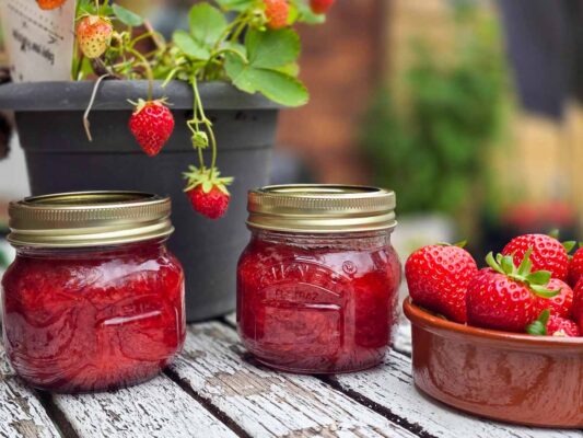 Two jars filled with strawberry jam, on a wooden table, outdoors. Behind there is a strawberry plant with a couple of red strawberries hanging. On the right there is a brown bowl filled with strawberries.