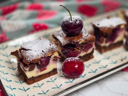 A rectangular platter with the square slices of cake on it. There are three slices. You can see the chocolate layer on the bottom, the white cheese one in the middle with the cherries on top, and the top chocolate layer. There are a few fresh cherries on the platter, and the cake has powder sugar sprinkled on the top.