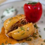 Romanian Vegan Stuffed Peppers Recipe – Ardei Umpluti de Post A close-up of two cooked stuffed peppers, a yellow one and a red one, on a white plate. One of the peppers is standing, whilst the other is cut in half, on the plate. The dish is sprinkled with chopped dill.