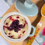 Romanian Rice Pudding Recipe – Orez Cu Lapte The rice pudding topped with sour cherry jam inside a blue bowl with handles. The lid of the bowl is leaning on the side.