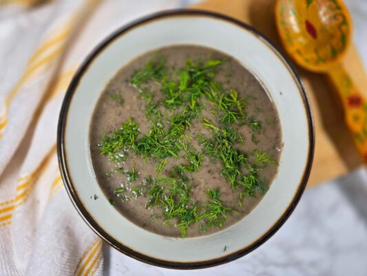 A white bowl with a golden rim filled with the brown soup, topped with chopped dill.