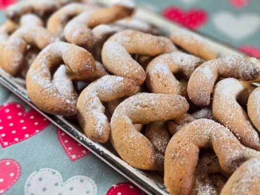 A rectangular platter filled with the walnut crescent cookies dusted with powdered sugar. It sits on a teal tea towel with pink and white hearts.