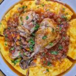 Romanian Chicken and Onion Stew Recipe – Tocanita de Ceapa cu Pui A grey plate with polenta on it. The chicken stew sits on top, with a lot of sauce. There is chopped parsley on top.
