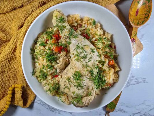 A deep white plate with the yellow pilaf in it, topped with a chicken leg and plenty of chopped dill. Around the bowl there is a yellow tea towel.