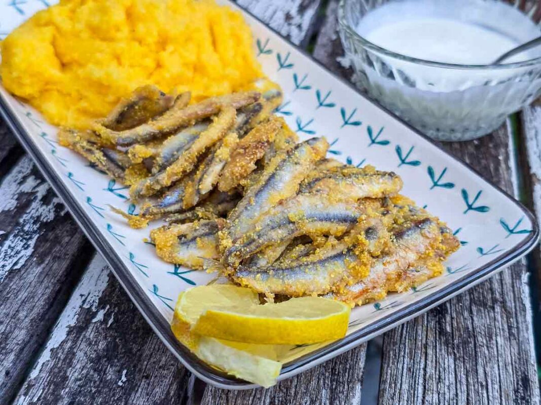 A rectangular platter with the fried fish on it, with a slice of lemon cut in half, and polenta in the back. Next to the platter there is a glass bowl filled with garlic sauce