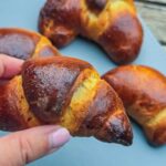 Romanian Chocolate Filled Crescent Rolls Recipe – Cornuri cu Ciocolata A close-up of my hand holding a chocolate filled crescent roll. Behind, you can see more rolls on a blue tray.