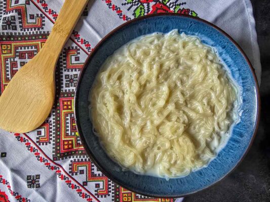 The milk soaked vermicelli inside a blue bowl, sat on a tea towel with Romanian patterns. Next to it there is a wooden spoon