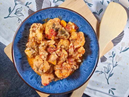 A blue bowl filled with the cauliflower and chicken stew. The bowl sits on a wooden board, next to a wooden spoon