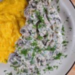 Romanian Garlic Mushrooms With Sour Cream Sauce Recipe – Ciuperci cu Smantana The garlic mushrooms with sauce on a gray plate, next to yellow polenta. There is chopped dill sprinkled on top.