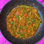 The Romanian green beans stew in a black pan, on an outdoors pink table.