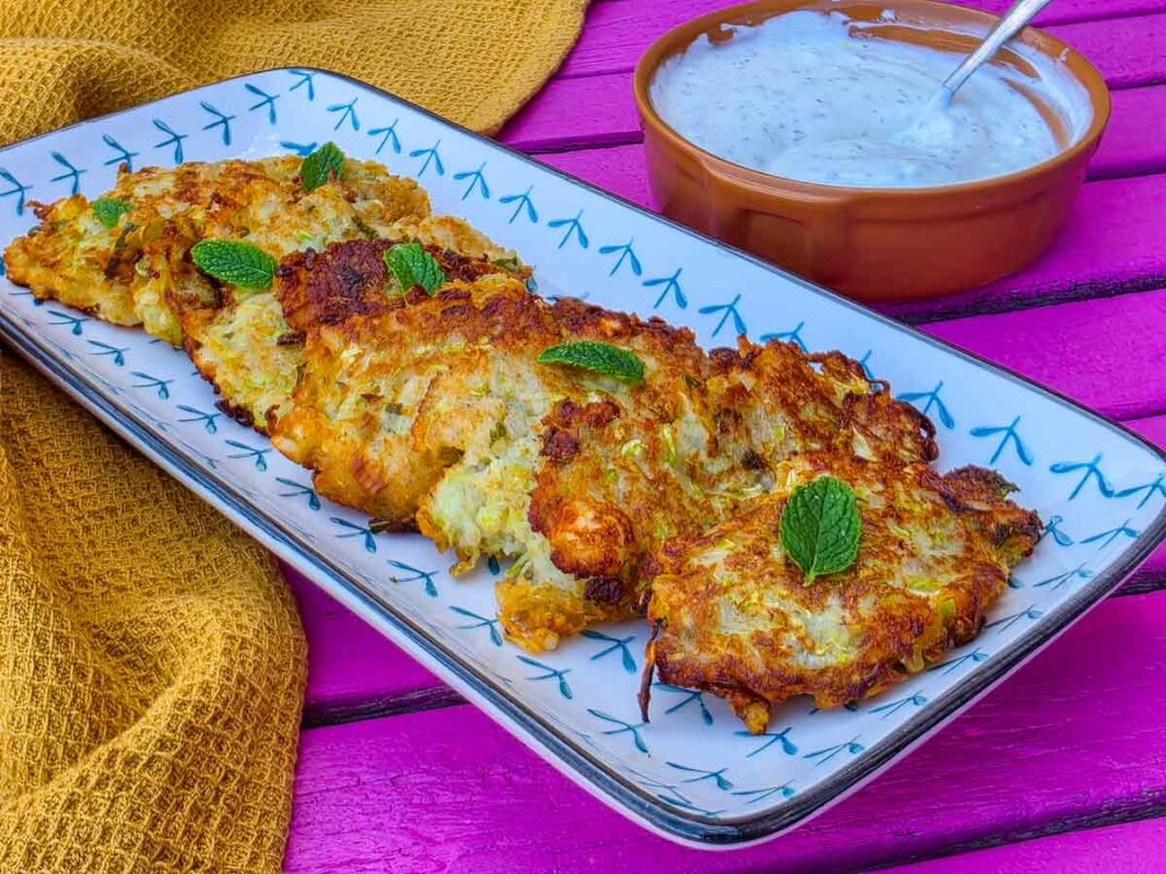 The fritters on a rectangular platter, topped with mint leaves. Next to it there is a yellow tea towel and a bowl with yogurt and garlic dip