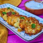 Romanian Zucchini Fritters With Cheese and Mint Recipe – Chiftele de Dovlecei cu Branza The fritters on a rectangular platter, topped with mint leaves. Next to it there is a yellow tea towel and a bowl with yogurt and garlic dip