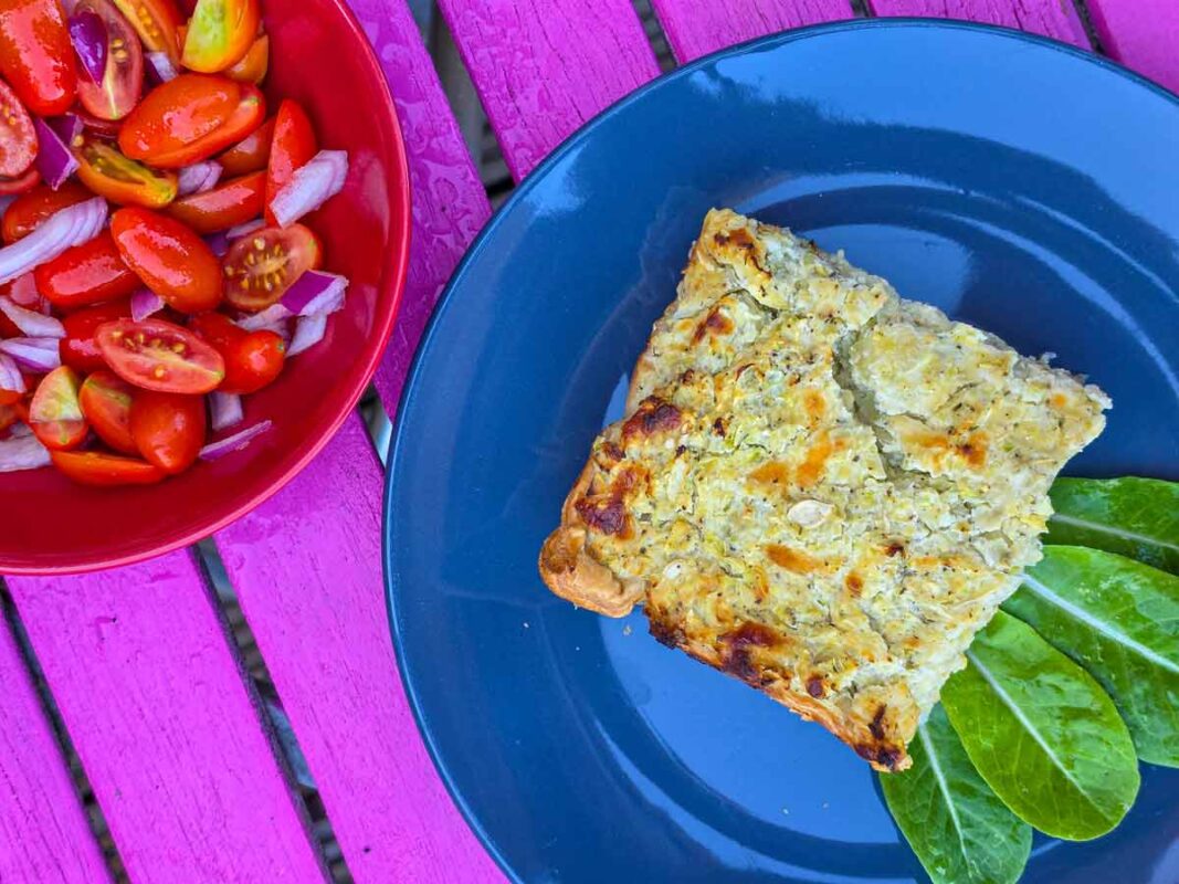 A close-up of the pie on a blue plate, next to a red bowl with a tomato salad inside. They are sitting on a pink outdoors table.