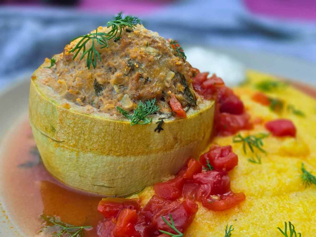 A close-up of a stuffed courgette next to polenta, with sour cream behind, and a red tomato sauce around.