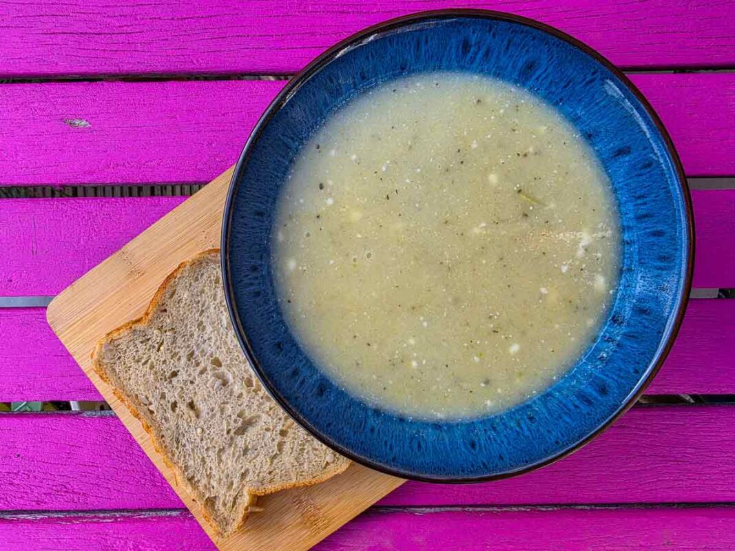 The soup inside a blue bowl, on a wooden board, with a slice of bread on the side. The table it sits on is pink
