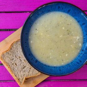 The soup inside a blue bowl, on a wooden board, with a slice of bread on the side. The table it sits on is pink